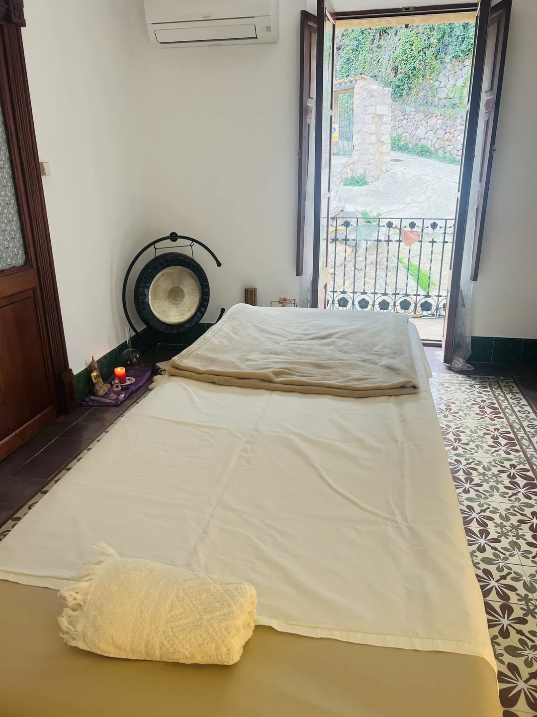 Ayurveda-Massageliege mit weißem Tuch und ein Gong in einem hellen Raum mit gemustertem Fliesenboden und Fenster ins Grüne, in Fornalutx.